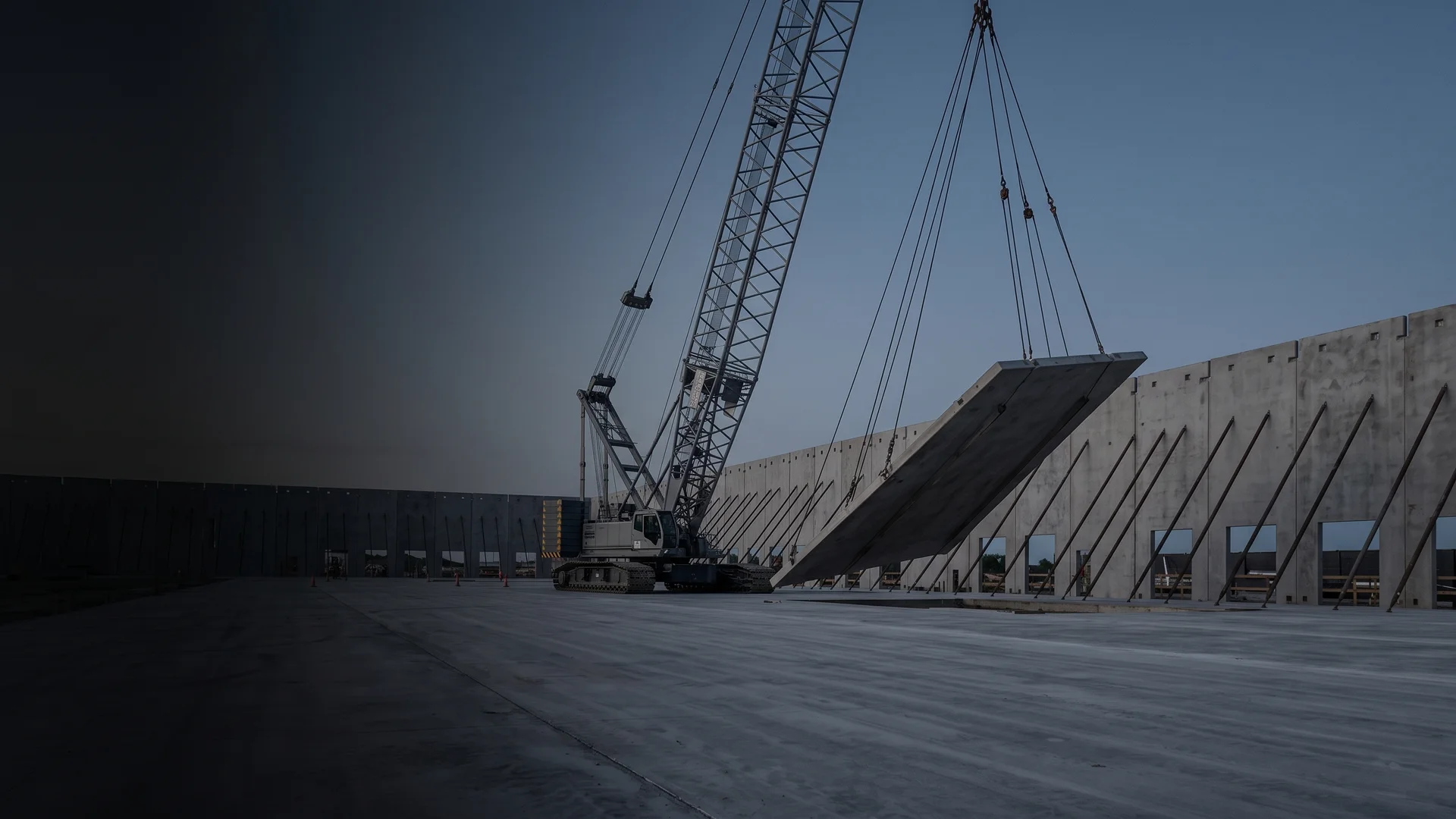 Crane tilting a concrete wall panel upright at dawn, braced tilt-wall panels behind it